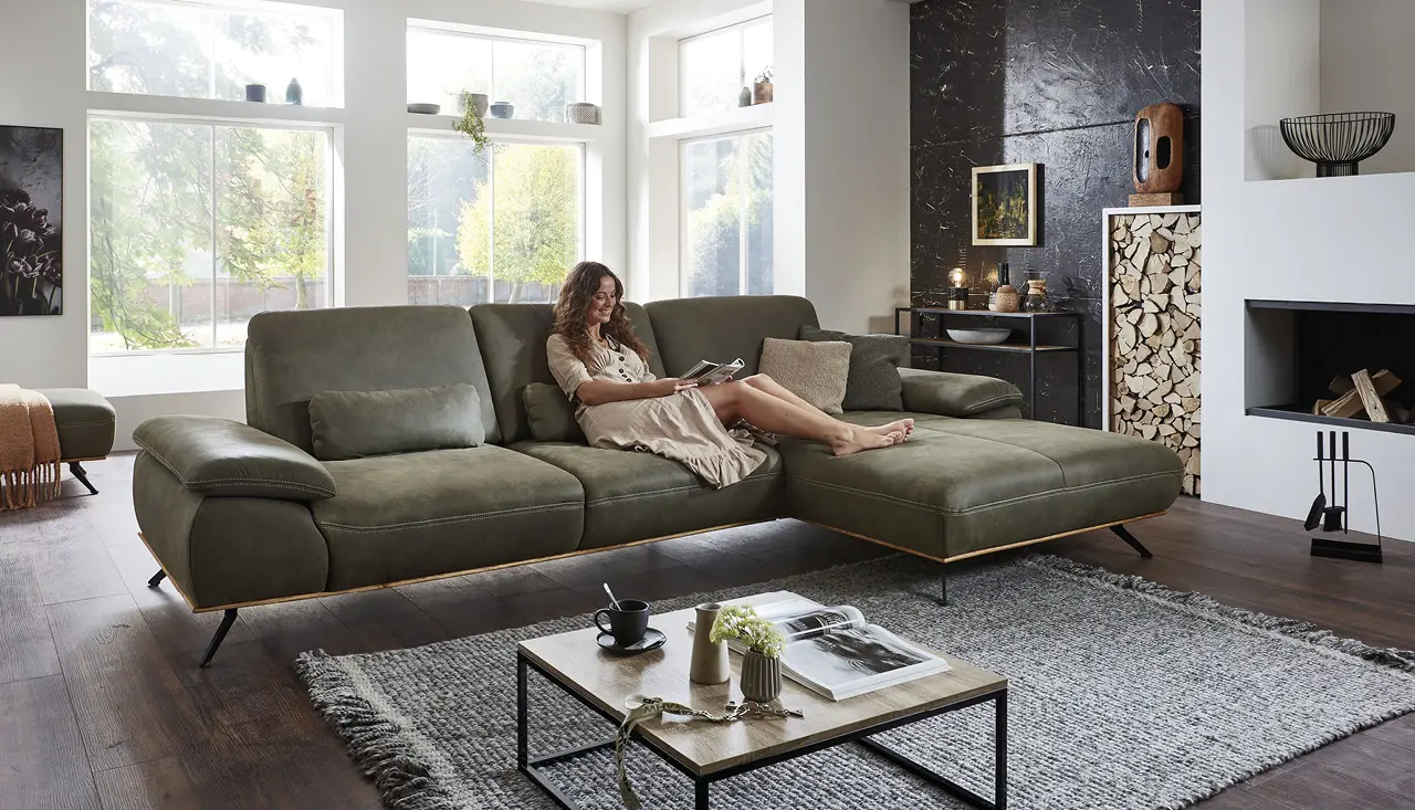 Frau entspannt auf modernem grünen Ecksofa im stilvollen Wohnzimmer mit Kamin, Holzdeko, Couchtisch und großer Fensterfront, gemütliche Wohnlandschaft im skandinavischen Design für behagliches Zuhause